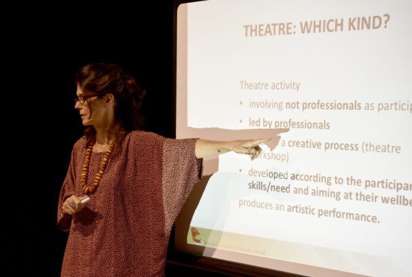 Alessandra Rossi Ghiglione on stage during the conference. She points with her finger to a detail on a slide showing the characteristics of community social theatre projected behind her.
