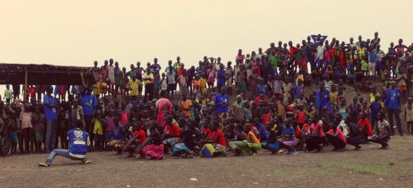 Wide-angle shot of the audience during an event in South Sudan. In the foreground, in front of the audience, South Sudanese men and women are crouched down, with bent knees, almost sitting on the ground, in rows of four one behind the other from left to right. In front of them is an IOM operator, also crouching like them. The audience watches the scene standing on a small hill.