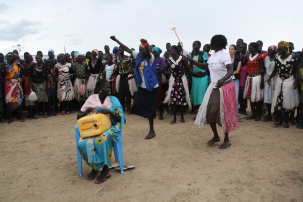 Three South Sudanese women perform in the center of a circle of people, both South Sudanese and others. Two of the women are standing and appear to be dancing; the third is sitting on a blue plastic chair between them and watching them, holding a yellow jerrycan resting on her legs.
