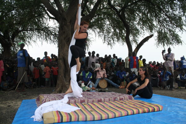 A performer, on the left, is preparing for aerial silks acrobatics; below her are mattresses. On the right, another girl, sitting on the ground, watches her. Around them is the local audience.