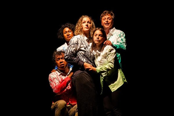 The ZID Theatre group on stage performing. Three women and two men, in white shirts with colorful red, blue, black, yellow, and green decorations (each person wearing one color). They are all close, huddled together as a group.