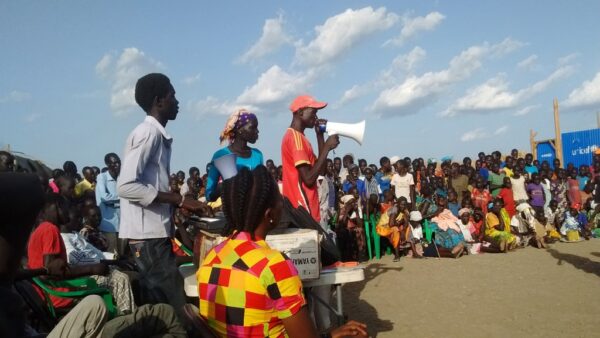 South Sudanese audience during a performance, some people sitting on the ground and others standing. In front of them, on the left, a small white table and two South Sudanese men and a woman standing, one of them holding a megaphone and speaking to the audience.