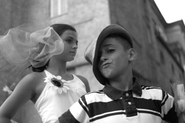 Black and white close-up of a boy and a girl during an event. The boy, wearing a striped shirt and a straw hat, looks at the camera playfully. The girl, behind him, wearing a tank top and a large fake sunflower on her chest and a tulle decoration in her hair, looks to the right