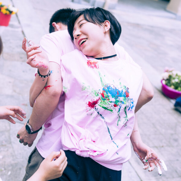 Close-up of two participants, back to back, with their arms intertwined. We only see her from the front, smiling, and him only from behind. Their shirts and hands are stained with paint of various colors.