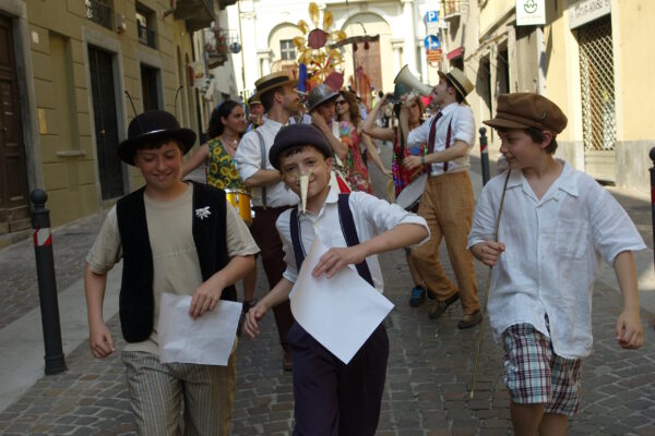 "Street parade. In the front row, children in costume, the one in the center has a fake Pinocchio nose. Behind them the SCT team, who animates the crowd with a megaphone, musicians, and many other participants. Some people, in the back, hold up large colorful paper constructions.