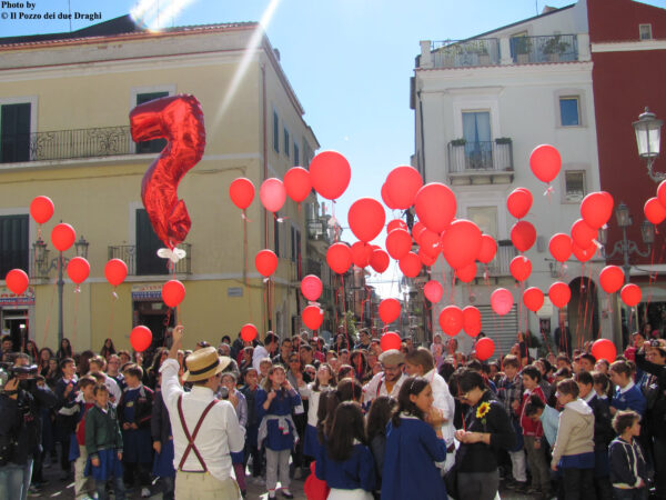 Large crowd in the square during an event, mainly boys and girls, together with the SCT team. The crowd holds red balloons