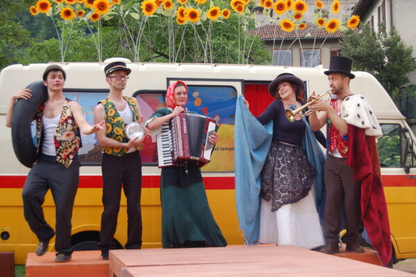 In front of the time post office, a wooden stage with performers on it. The three men wear vests with very colorful patterns and hats. The women wear long dresses. The performers play, accordion and trumpet, and dance