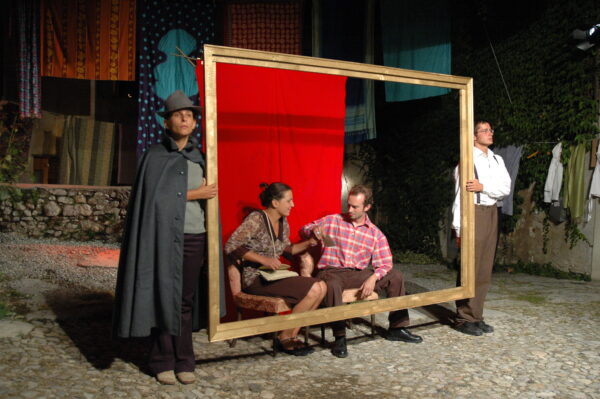 Actors performing. Two hold a very large golden frame. Behind the frame we see an actor and an actress, sitting on two armchairs, giving the illusion of being framed. Behind them a red backdrop.