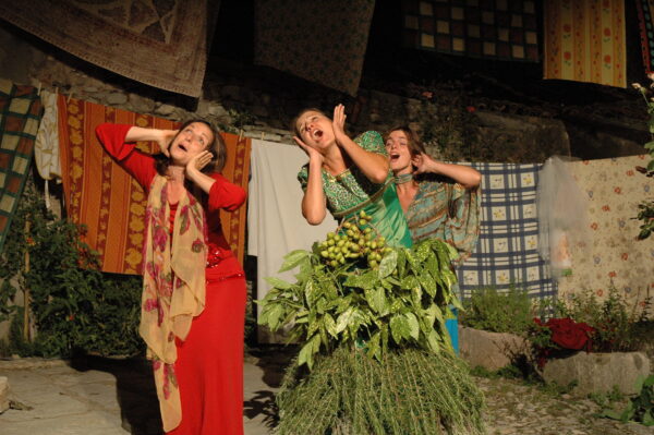 Three women performing amidst colorful hanging laundry. One is dressed in red, with a beige scarf with red flowers around her neck, one is dressed in green and blue, with a patterned shirt, one is dressed in green, with a gold-decorated bodice. In the middle of them a structure of herbs, branches and leaves.