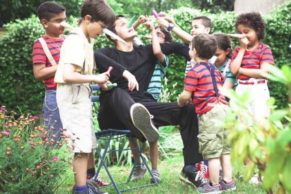 In a meadow, among flowers and hedges, an SCT worker sitting on a chair, dressed in black, with a paper Pinocchio nose. Around him are seven boys and girls, with the same nose, dressed as Pinocchio, playing with him.