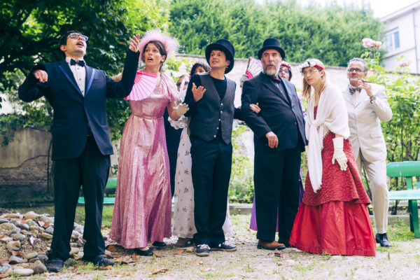 A group performing. Five men in elegant suits, black or beige; four women in vintage dresses, colorful, with fans in their hands.