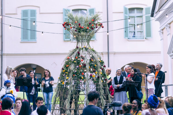 A courtyard full of people, some on stage with lecterns and microphones, others in the audience. In the center, an hourglass-shaped structure made of intertwined twigs, herbs, plants and flowers.