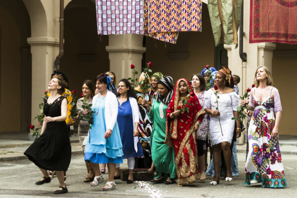 A group of women, of different cultures and ages, dressed in traditional, elegant and colorful clothes, hold roses and walk in a compact group, inside a courtyard.