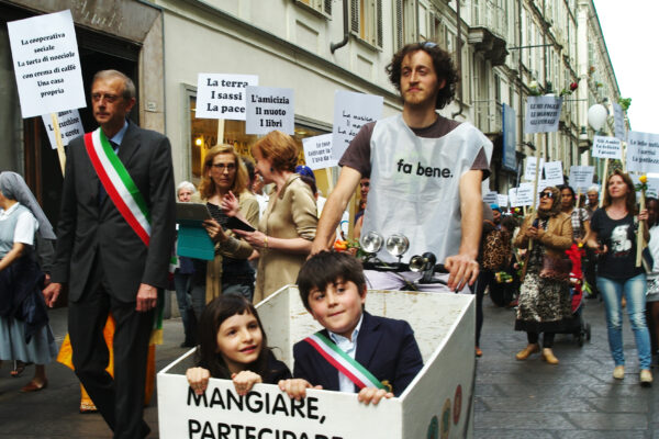 Crowd in the street during a community parade. People are holding signs. In the foreground, a boy pushes a cart with a boy and a girl inside.