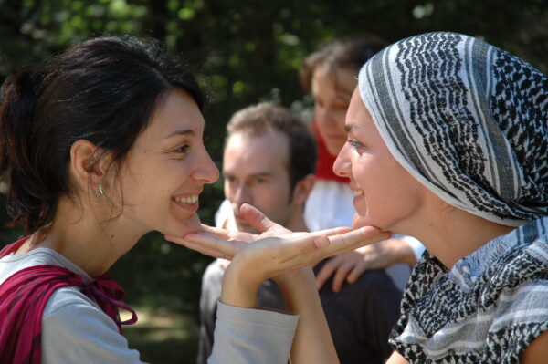 Close-up of two girls, one of whom has her head covered by a scarf. The girls are facing each other, looking into each other's eyes and smiling, and they both have one hand, palm up, under the other's chin