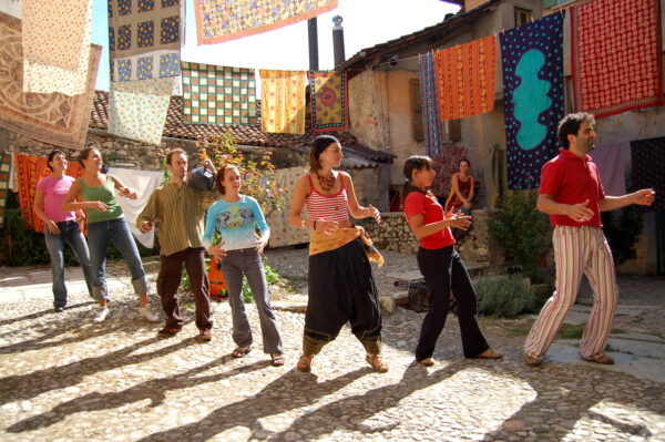 Group of performers dancing in an internal courtyard, where there are many clothes hanging on high wires, all colorful and with cheerful patterns