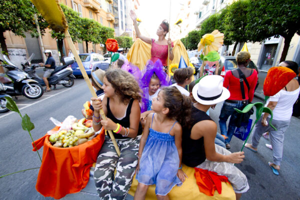 A float through the city streets, on which there are boys and girls and a woman. They are dressed in colorful clothes, with lots of tulle and decorations, and they hold large papier-mâché flowers. On the float there is also a basket with a lot of fruit collected for the Community Macedonia. Other participants walk near the float.