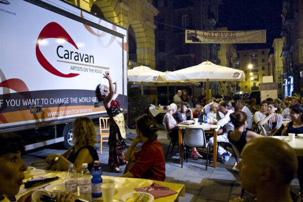 The photo was taken in the evening. The caravan used for the project is parked in a small square, surrounded by tables from a bar. Many people are sitting at the tables, eating and watching the Caravan performer, standing among them, who is performing