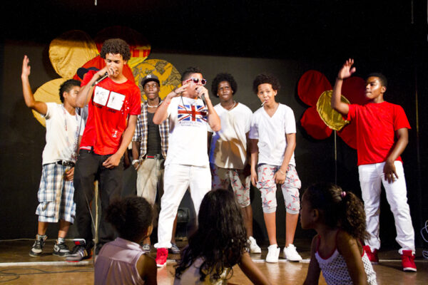 Group of seven young poc boys on stage. Two of them are rapping. In front of them, three little girls are sitting on the ground watching them