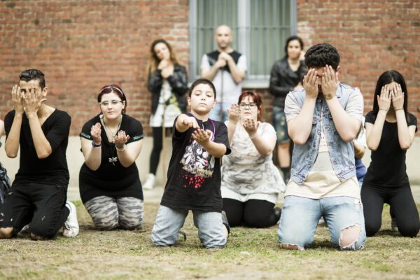 A group of young people performing, kneeling on the ground in a meadow, covering their faces with their hands.