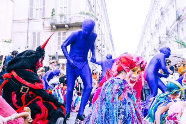 Performers in the city streets, dressed in blue bodysuits that cover their entire body and head, or with colorful Carnival-style masks. Around them are participants and onlookers.