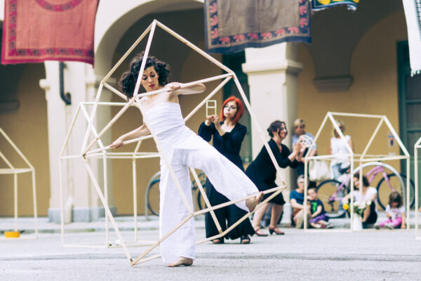 In a courtyard, a woman dances with a large wooden structure resembling a stylized house, expressing movement and grace. Behind her, other identical structures, and two other women perform interacting with smaller structures.