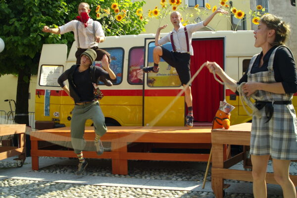 In front of the time post office, a wooden stage on which two performers in shirts and suspenders dance. Below the stage other performers, with a rope in hand, spin it while one of them jumps.