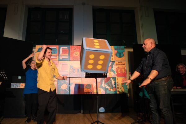 Un attore e un'attrice sul palco con un uomo del pubblico che lancia il grande dado di cartone azzurro e giallo. Dietro di loro le caselle del gioco dell'oca