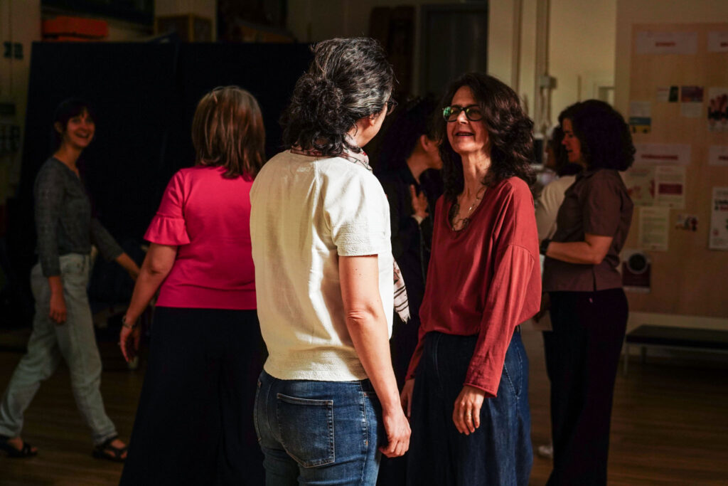 Due partecipanti si confrontano durante un’attività teatrale di gruppo, mentre altre persone lavorano in coppia sullo sfondo.